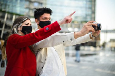 Covid ya da Coronavirus maskeli mutlu turistler şehirde birlikte yürüyüp ilginç bir noktayı fotoğraflıyorlar.