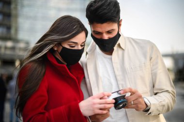 Covid ya da Coronavirus maskeli mutlu turistler bir şehirde birlikte yürüyüp birlikte çekilen fotoğraflara bakıyorlar.