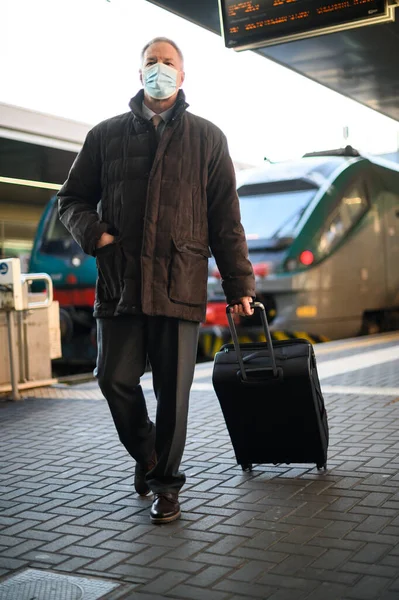 シニア旅行者は 彼のトロリーと電車の駅を歩くとコロナウイルスのパンデミックに対する保護マスクを着て — ストック写真