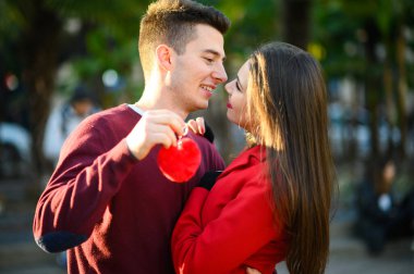 Genç bir adam elinde kalp şeklinde bir kolye tutarken kız arkadaşını öpüyor.