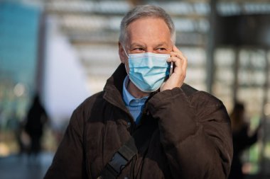 Covid Coronavirus konsepti, maskeli yaşlı iş adamı dışarıda akıllı telefonuyla konuşuyor.