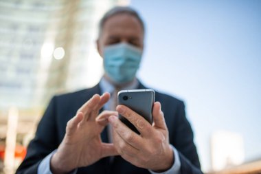 Covid konsepti, maskeli yaşlı iş adamı akıllı telefonunu açık havada bir şehirde kullanıyor.