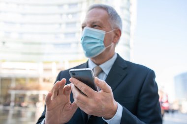 Covid konsepti, maskeli yaşlı iş adamı akıllı telefonunu açık havada bir şehirde kullanıyor.