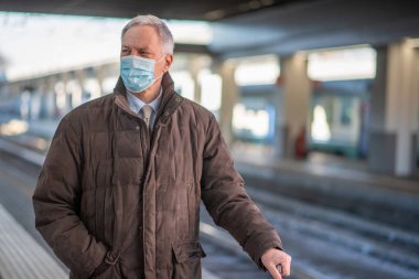 Maskeli kıdemli işadamı treni bekliyor, covid ve coronavirüs hareketlilik konsepti