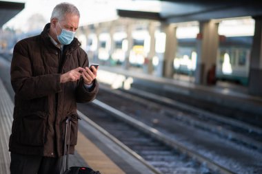 Maskeli kıdemli işadamı trenini beklerken akıllı telefonunu kullanıyor. Covid ve Coronavirus hareketlilik konsepti.