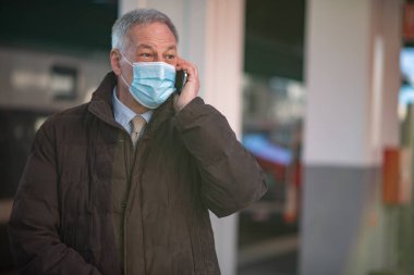 Covid Coronavirus konsepti, maskeli yaşlı işadamı tren istasyonunda akıllı telefonuyla konuşuyor.