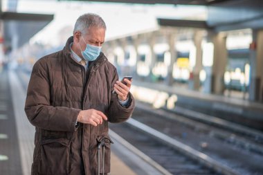 Maskeli kıdemli işadamı trenini beklerken akıllı telefonunu kullanıyor. Covid ve Coronavirus hareketlilik konsepti.