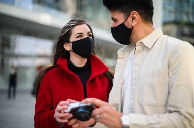 Covid ya da Coronavirus maskeli mutlu turistler bir şehirde birlikte yürüyüp birlikte çekilen fotoğraflara bakıyorlar.