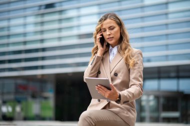 Dışarıda tablet tutarken cep telefonunu kullanan genç bir iş kadını.