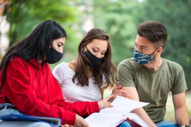 Üç öğrenci, Coronavirus zamanlarında bankta oturup maske takarak ders çalışıyor.