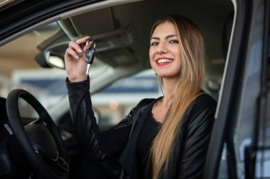 Genç ve mutlu bir kadın yeni arabasının anahtarını bir araba galerisinde gösteriyor.