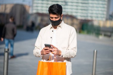Akıllı telefon, kovid ve koronavirüs konsepti kullanarak alışveriş çantası taşıyan maskeli bir adam