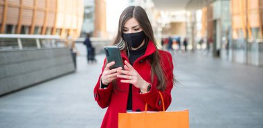 Cep telefonunu kullanarak şehirde dolaşan maskeli genç bir kadın.