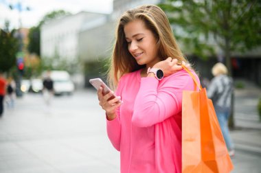 Şehirde alışveriş yaparken akıllı telefonunu kullanan genç bir kadın.