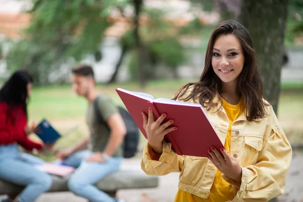 Woman student holding a book in front a a group of friends in park