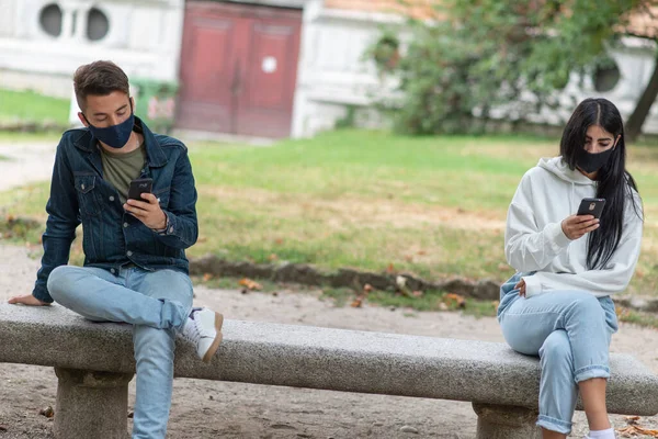 Young people keeping social distancing due to coronavirus while using their phones outdoor in a park