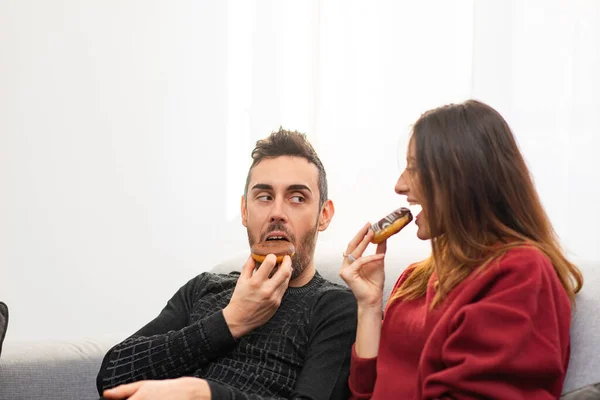 幸せなカップルボーイフレンドとガールフレンドや妻と夫食べるドーナツで彼らのアパートのリビングルーム — ストック写真