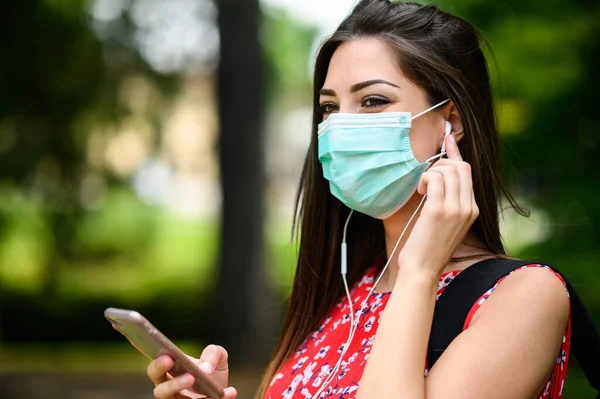 女性学生が公園を歩きながらスマートフォンを使って音楽を聴いたり マスクをしてコロナウイルスから身を守る Covid 19パンデミック — ストック写真