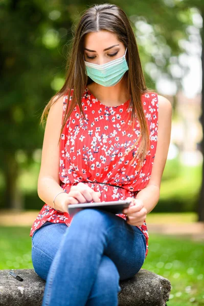 公園のベンチに座ってマスクを着用するデジタルタブレットを使用して若い女性 コロナウイルスの概念 — ストック写真