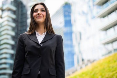 Çim tarlasının yanında gülümseyen genç bir kadın ve modern bir şehirde cam ofis binası.