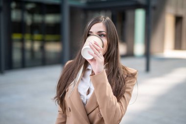 Genç iş kadını şehirde yürürken kahve içiyor.