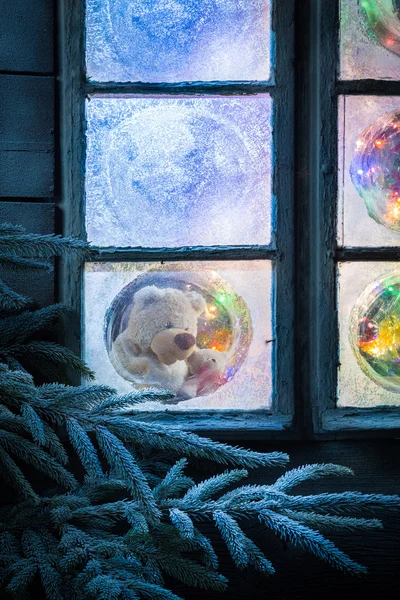 Urso de pelúcia na janela congelada para o Natal com árvore e luzes — Fotografia de Stock