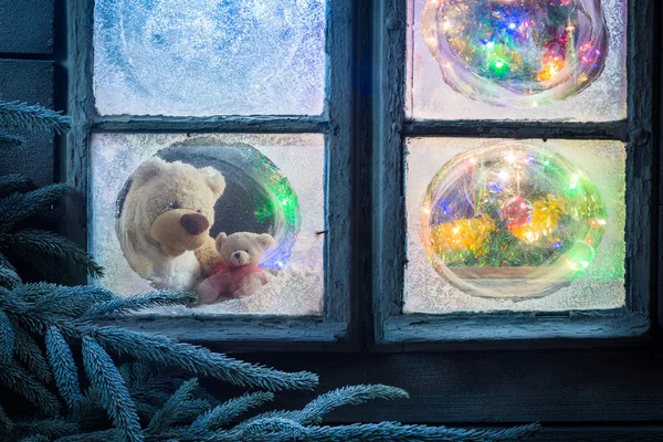 Teddy bear for Christmas in frozen window with tree and lights — Stock Photo, Image