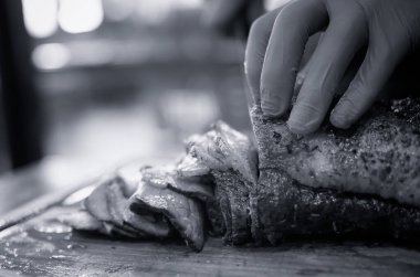 Ahşap tahtada ızgara ve az pişmiş biftek dilimleyen şeflerin eli kapalı. Siyah ve beyaz ton.