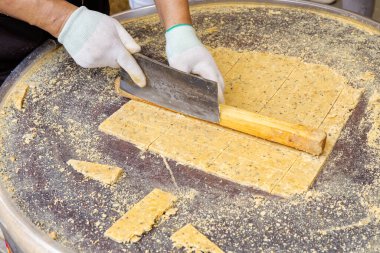 Bir sokak satıcısının ya da fırıncının yakın çekimi büyük bir tabak geleneksel sarı fıstık şekeri ya da susamlı aperatifi yuvarlak metal bir masada büyük bir bıçakla kesiyor. Eller eldiven giyer, şeker yapma sürecini veya Asya sokak yemeklerini betimler.