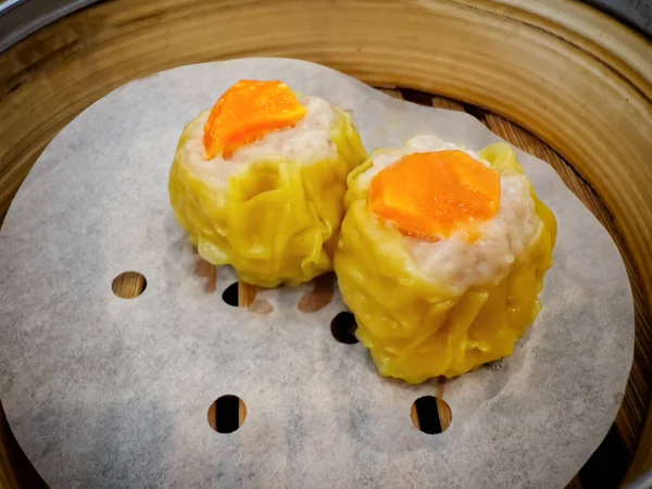 Parşömen kağıdının üzerinde geleneksel bambu budan bir buharlı tencerede servis edilen parlak turuncu tuzlu yumurta sarısı ile kaplanmış Shumai adlı iki domuz etli böreğinin yakın plan çekimi.