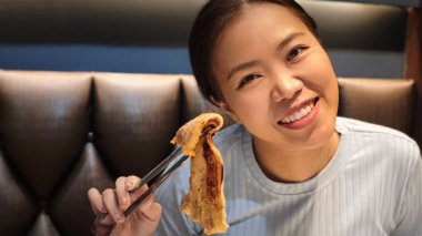 Açık mavi tişörtlü neşeli bir Asyalı kadın elinde metal maşayla sulu ızgara domuz göbeği tutarken kameraya gülümsüyor.