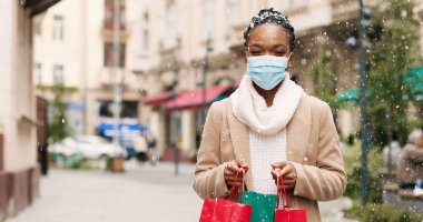Kar yağışlı kış şehrinde maskeli güzel mutlu Afro-Amerikan kadın Noel arifesinden önce alışveriş yaptıktan sonra Noel hediyeleriyle. Dışarıdan hediye getiren çantalı kadın portresi. Tatil sezonu konsepti