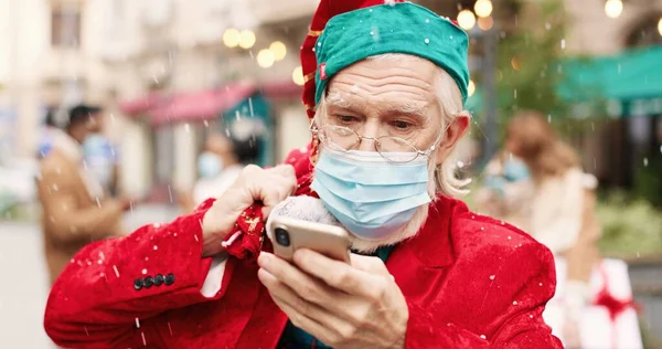 Close up portrait of old Santa Clause in mask and glasses texting on cellphone while standing in snowy crowded street and holds bag full of gifts. Christmas spirit. Santa tapping on smartphone.