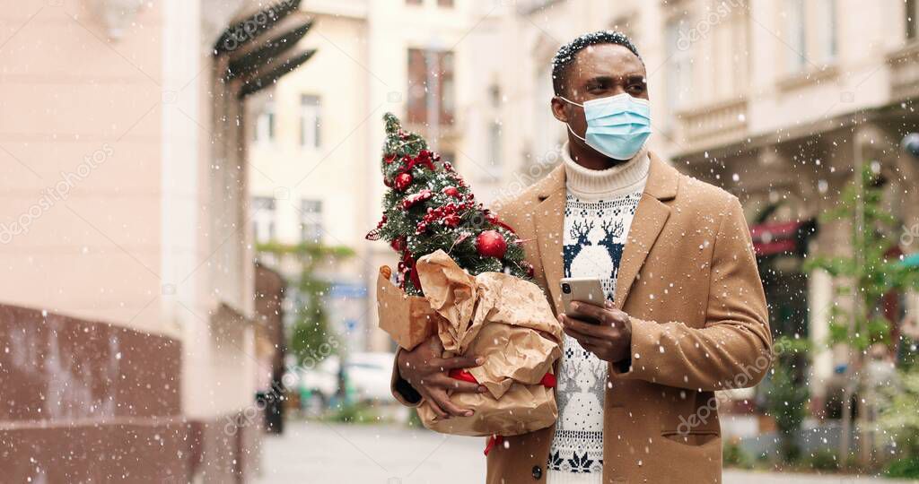 Close up portrait of happy African American man in medical mask standing in snowy town and typing on smartphone. Handsome male with little new year tree outdoor and texting on cellphone. Christmas