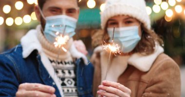Karantinadaki tıbbi maskelerle süslenmiş Xmas caddesinde dururken ellerinde maytap olan beyaz mutlu çiftin yakın çekimleri. 2021 yılını kutlayan neşeli adam ve kadın. Tatil kutlaması konsepti