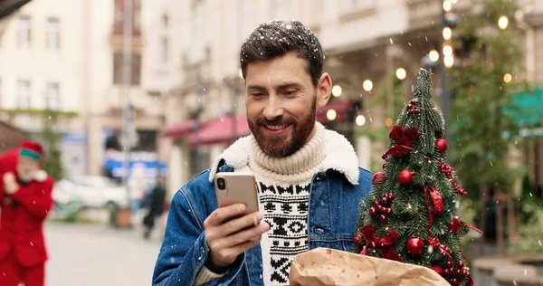 Handsome joyful Caucasian man standing in decorated crowded city and texting on smartphone and receiving holiday greetings. Close up portrait of male with small christmas tree typing on cellphone