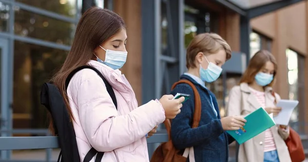 Porträtt av tonåriga vita skolbarn som står utomhus nära skolan och läser. Koncentrerad vacker flicka i mask läser ur copybook. Pojke med planerare innan lektionerna. Höstkoncept — Stockfoto