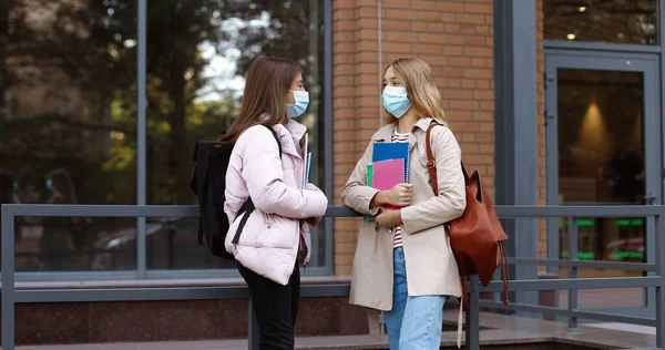 Vackra unga tonårstjejer i masker med ryggsäckar som står nära skolan och pratar. Kaukasiska skolpojke hälsning med armbågar med glada kvinnliga elever med copybooks utomhus. Karantänkoncept — Stockfoto