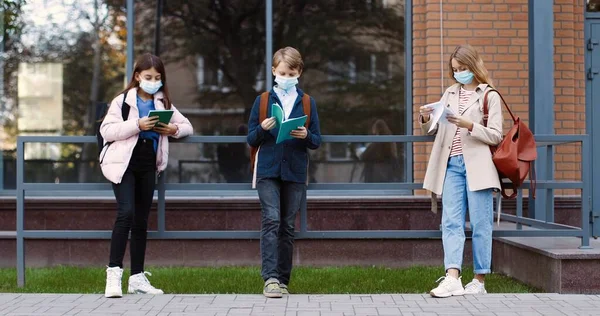 Kaukasiska tonårselever står utomhus nära skolan och läser ur anteckningsböcker. Koncentrerade flickor i masker och pojke med copybooks lärande före examen. Studera inför klasser Utbildningskoncept — Stockfoto