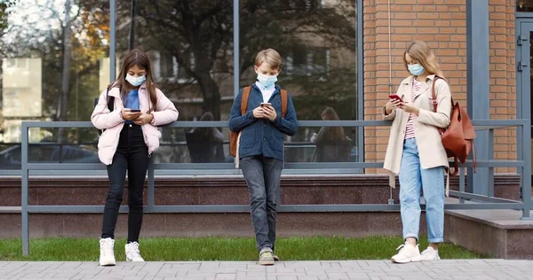 Studenti caucasici adolescenti che stanno all'aperto vicino alla scuola e leggono dai quaderni. Ragazze concentrate in maschera e ragazzo con copybook imparare prima dell'esame. Studiare prima delle lezioni Concetto educativo — Foto Stock
