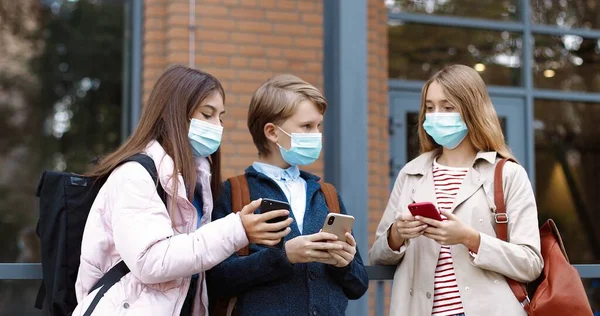 Porträtt av vita tonåringar som står nära skolan och skriver på smartphones. Vackra flickor i masker pratar och visar pojken något på mobilen. Klasskamrater med ryggsäckar Skolkoncept — Stockfoto