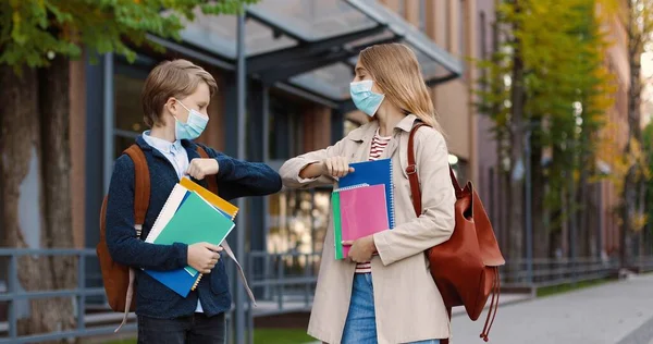 Porträtt av tonåring pojke i mask med ryggsäck möte nära skolan med flicka klasskamrat och prata. Vit skolflicka hälsning med armbåge med vän med copybooks och anteckningsböcker utomhus Studiekoncept — Stockfoto