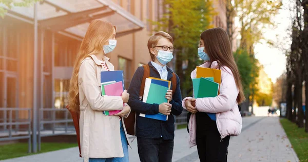 Porträtt av glada vita skolbarn som står utomhus och pratar efter lektionerna. Glada vackra tonårsflickor i masker med copybooks och pojke i glasögon chattar på skolgården Utbildningskoncept — Stockfoto