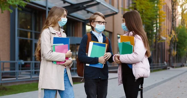 Porträtt av glada vita skolbarn som står utomhus och pratar efter lektionerna. Glada vackra tonårsflickor i masker med copybooks och pojke i glasögon chattar på skolgården Utbildningskoncept — Stockfoto