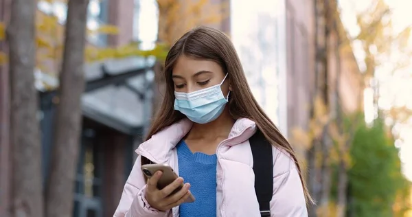 Nahaufnahme Porträt eines jungen Teenie-Mädchens in Maske, das mit Rucksack und SMS auf dem Handy draußen auf der Straße spaziert. Kaukasisch schöne Schülerin nach dem Unterricht tippt auf dem Smartphone. Quarantäne-Konzept — Stockfoto