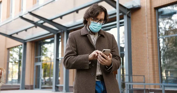 Porträtt av vit vacker manlig lärare i glasögon och mask knacka och skriva på smartphone utomhus. Flickor och pojkar skolelever med copybooks körs till pedagog och ställa frågor — Stockfoto