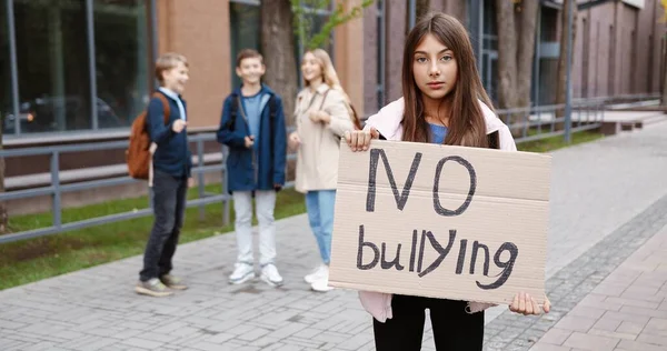 Retrato da jovem colegial caucasiana em pé ao ar livre com sinal de "Sem bullying", enquanto as crianças rindo dela no fundo. Linda estudante segurando cartão contra zombar. Conceito escolar — Fotografia de Stock