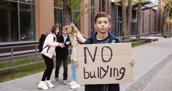 Porträt eines fröhlichen kaukasischen Teenagers, der mit einem Schild "Kein Mobbing" im Freien steht, während Teenager ihn im Hintergrund auslachen. Männlich glücklicher Schüler in der Nähe der Schule gegen Spott. Konzept zur sozialen Frage — Stockfoto