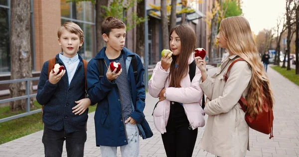 Retrato de un joven estudiante caucásico feliz parado al aire libre y hablando mientras come manzanas. Niños y niñas alumnos con mochilas comiendo bocadillos cerca de la escuela en la calle. Concepto Amigos — Foto de Stock