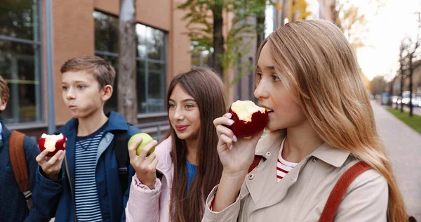 Sluiten van vrolijke blanke scholieren die buiten appels eten. Jongens en meisjes schoolvrienden in goed humeur hebben een snack bij de lunch in de buurt van school in goed humeur. Voedselconcept — Stockfoto